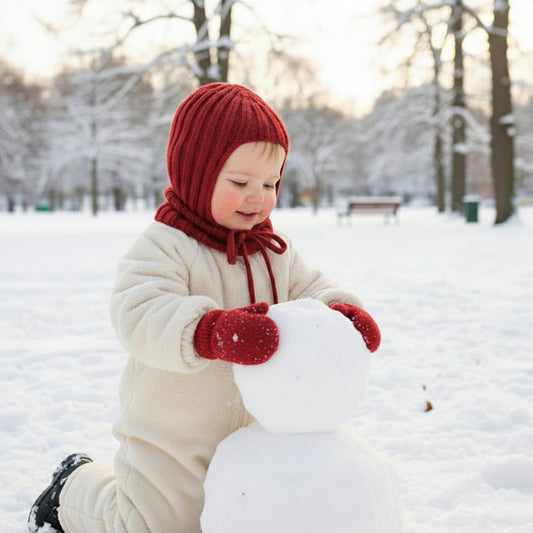 child wearing a red helmet hat and matching mittens building a snowman in a park
